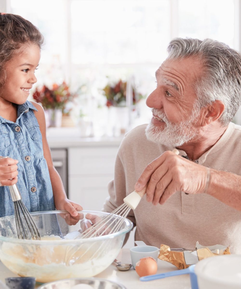 Young girl and grandfather making cake mixture at the kitchen table, smiling at each other, close up